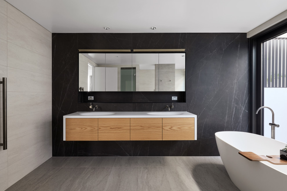 This is an example of a contemporary bathroom in Perth with flat-panel cabinets, medium wood cabinets, a freestanding tub, black tile, an integrated sink, grey floor, white benchtops, a double vanity and a floating vanity.