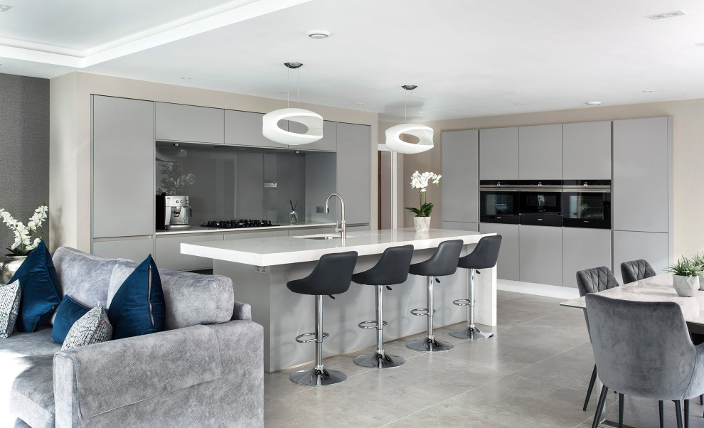 This is an example of a large contemporary l-shaped eat-in kitchen in Belfast with flat-panel cabinets, granite benchtops, with island, an undermount sink, grey cabinets, grey splashback, glass sheet splashback, porcelain floors, grey floor and white benchtop.
