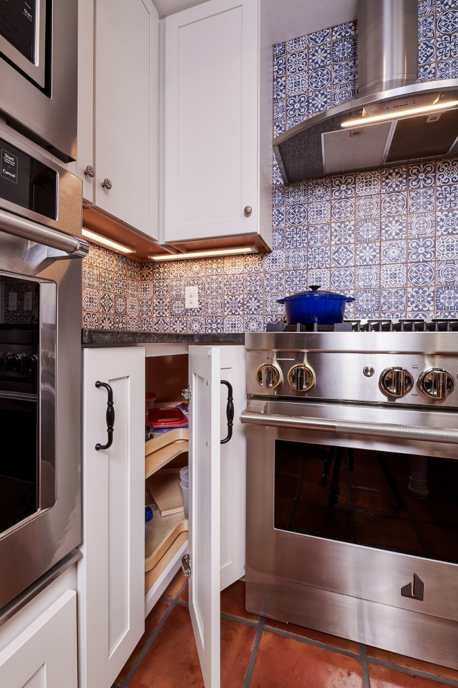 他の地域にある高級な中くらいなサンタフェスタイルのおしゃれなキッチン (エプロンフロントシンク、シェーカースタイル扉のキャビネット、白いキャビネット、御影石カウンター、青いキッチンパネル、テラコッタタイルのキッチンパネル、シルバーの調理設備、テラコッタタイルの床、赤い床、茶色いキッチンカウンター、折り上げ天井) の写真