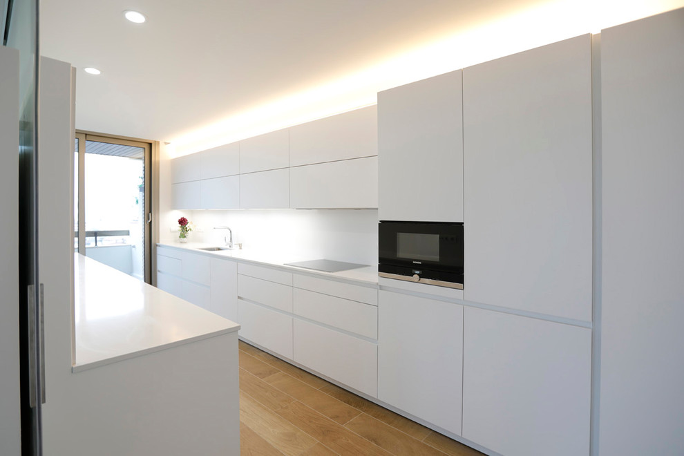 Modern galley kitchen in Other with flat-panel cabinets, white cabinets, black appliances, a single-bowl sink, white splashback, light hardwood floors and white benchtop.