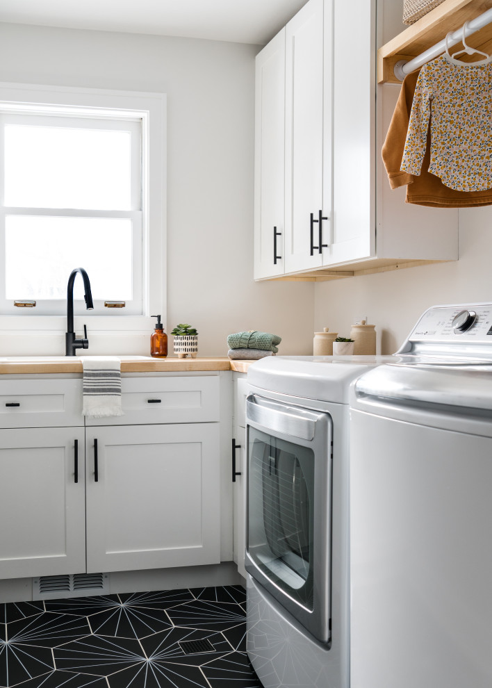 Photo of a small midcentury galley dedicated laundry room in Chicago with a drop-in sink, shaker cabinets, white cabinets, wood benchtops, white walls, ceramic floors, a side-by-side washer and dryer, black floor and beige benchtop.