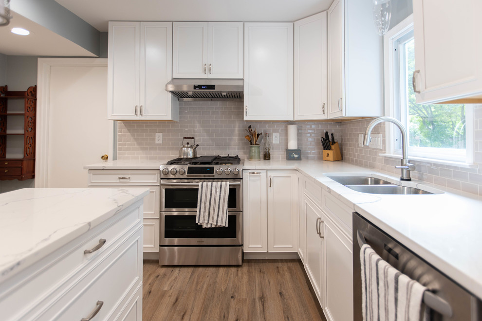 Large contemporary u-shaped open plan kitchen in DC Metro with an undermount sink, raised-panel cabinets, white cabinets, quartz benchtops, grey splashback, subway tile splashback, stainless steel appliances, vinyl floors, with island, brown floor and white benchtop.