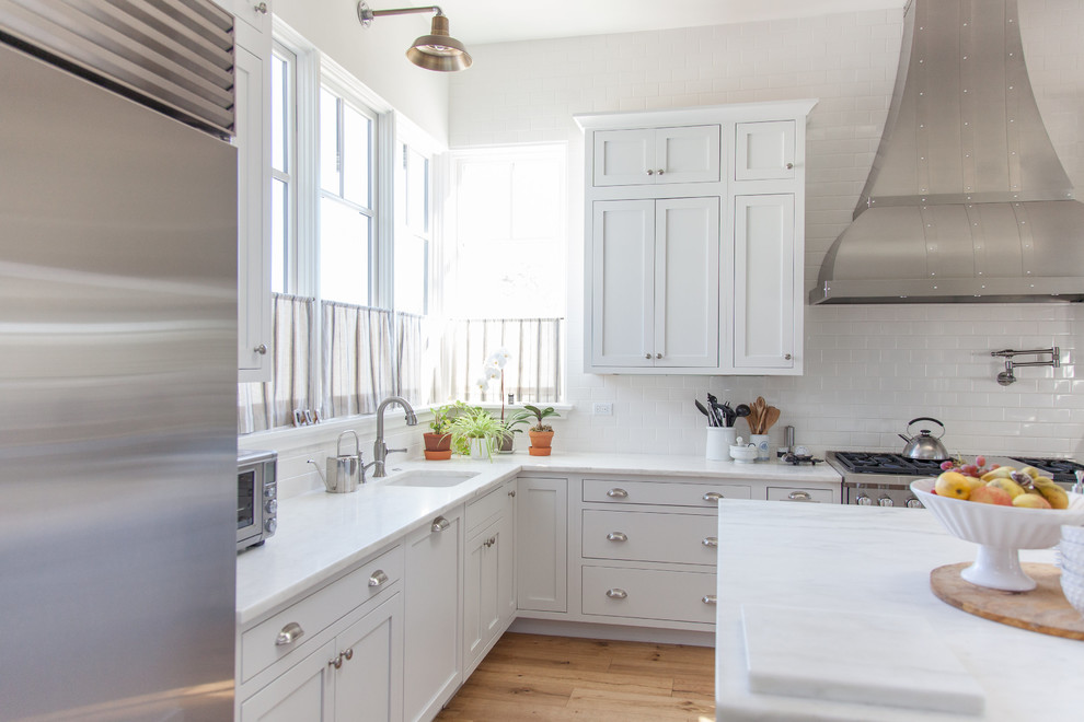 Large transitional l-shaped light wood floor eat-in kitchen photo in Phoenix with a farmhouse sink, white cabinets, white backsplash, stainless steel appliances, an island, shaker cabinets, stainless steel countertops and porcelain backsplash