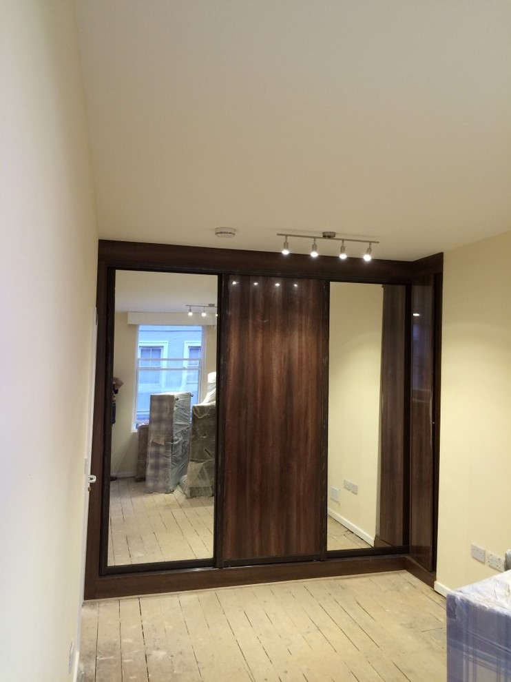 Photo of a mid-sized contemporary gender-neutral built-in wardrobe in London with glass-front cabinets, dark wood cabinets and light hardwood floors.