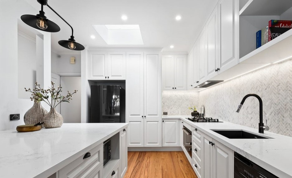 Inredning av ett klassiskt mellanstort vit vitt kök, med en undermonterad diskho, luckor med upphöjd panel, vita skåp, bänkskiva i kvarts, flerfärgad stänkskydd, stänkskydd i keramik, svarta vitvaror, mörkt trägolv, en köksö och brunt golv