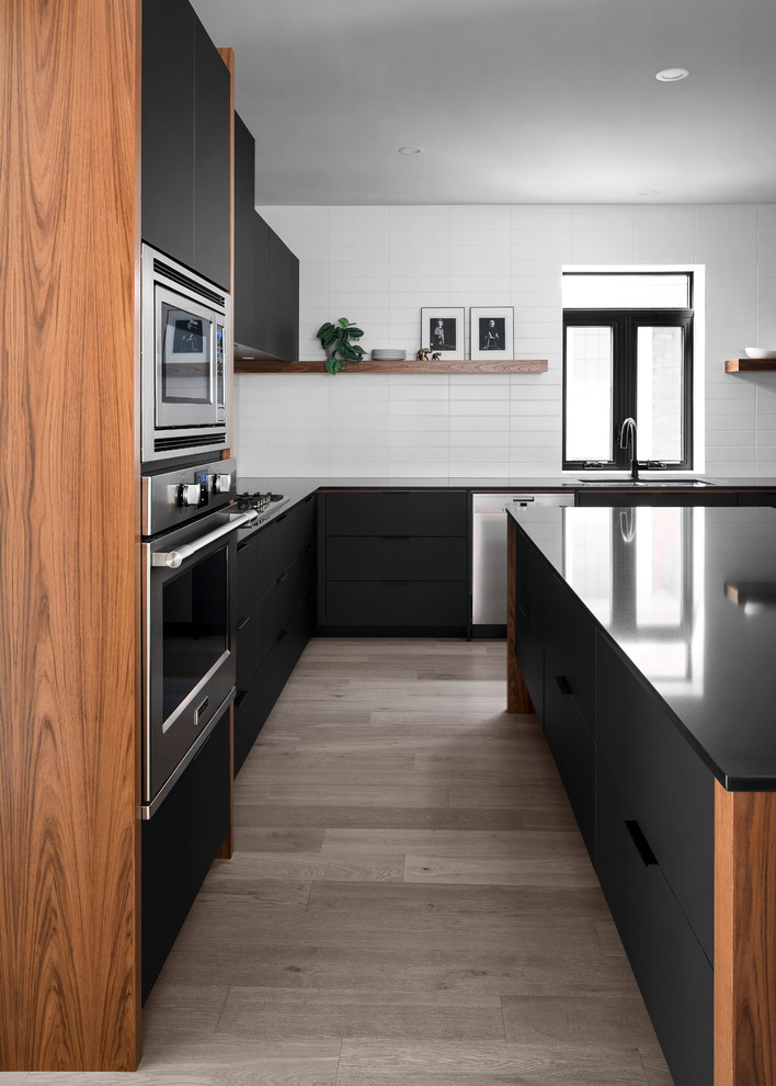Mid-sized midcentury u-shaped eat-in kitchen in Toronto with an undermount sink, flat-panel cabinets, black cabinets, quartz benchtops, white splashback, subway tile splashback, stainless steel appliances, light hardwood floors, with island, brown floor and black benchtop.