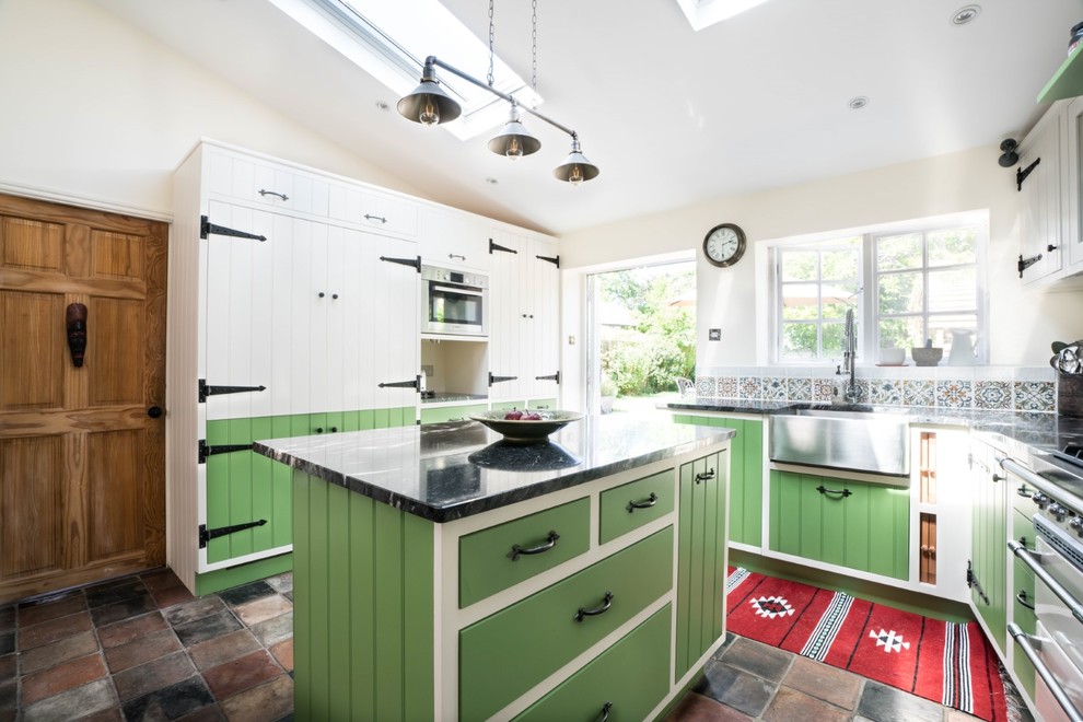 This is an example of a small country l-shaped eat-in kitchen in Essex with a farmhouse sink, green cabinets, granite benchtops, multi-coloured splashback, mosaic tile splashback, panelled appliances, porcelain floors, with island, multi-coloured floor and black benchtop.