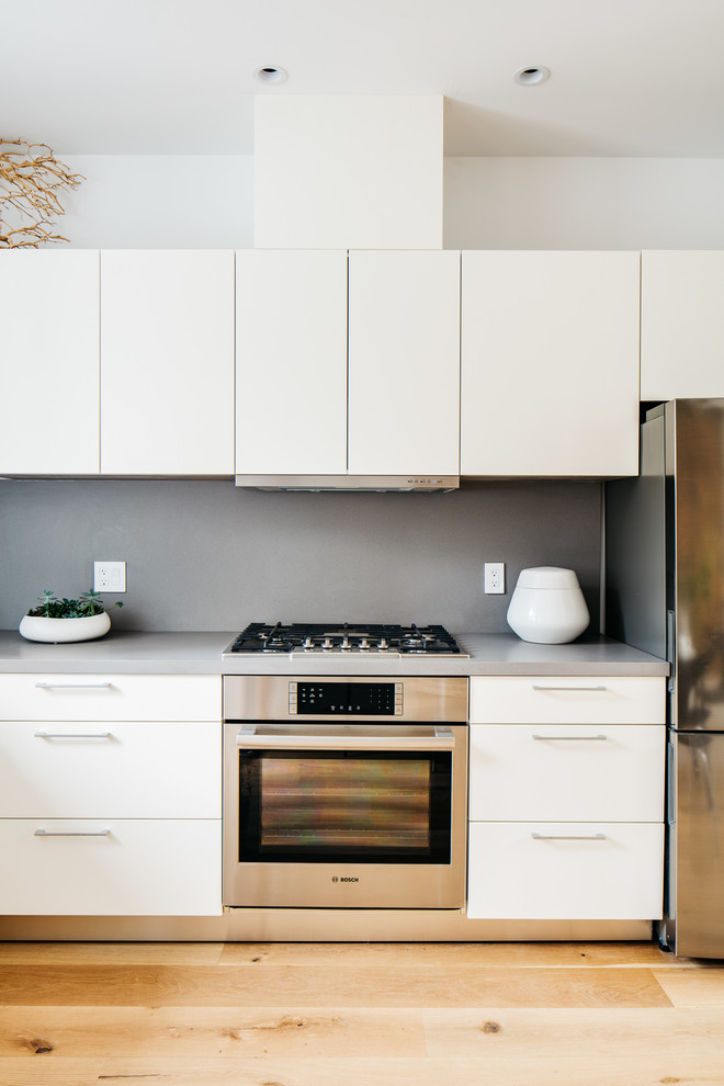 This is an example of a mid-sized modern l-shaped eat-in kitchen in San Francisco with an undermount sink, flat-panel cabinets, white cabinets, solid surface benchtops, grey splashback, stone slab splashback, stainless steel appliances, light hardwood floors, no island and grey benchtop.