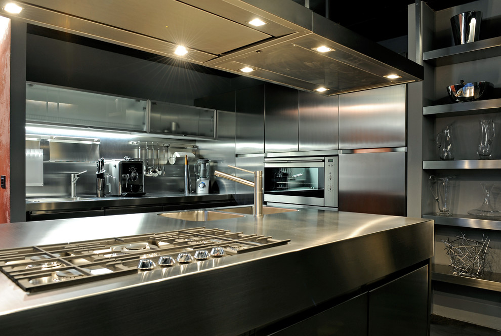 This is an example of an industrial kitchen in Milan with an integrated sink, flat-panel cabinets, stainless steel cabinets, stainless steel benchtops, metallic splashback, metal splashback and stainless steel appliances.