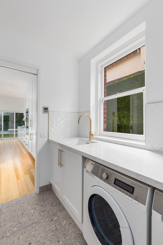 Large modern galley laundry room in Melbourne with an undermount sink, shaker cabinets, quartz benchtops, white benchtop, white cabinets, white splashback, subway tile splashback, porcelain floors and a side-by-side washer and dryer.