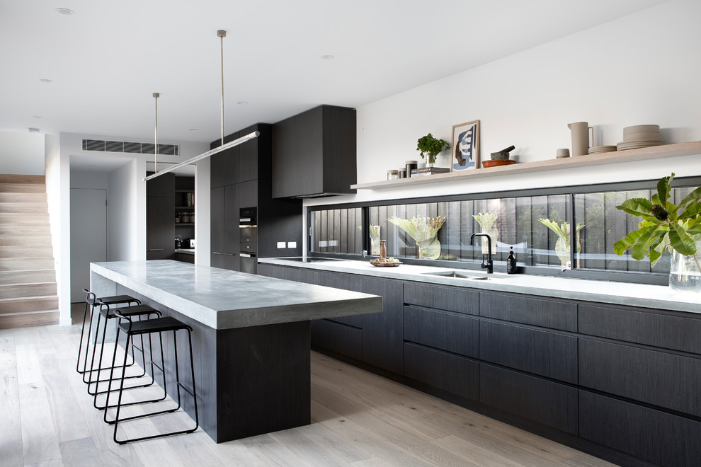 Photo of a modern galley kitchen in Melbourne with a double-bowl sink, concrete benchtops, light hardwood floors, with island, flat-panel cabinets, black cabinets, window splashback, black appliances, beige floor and grey benchtop.