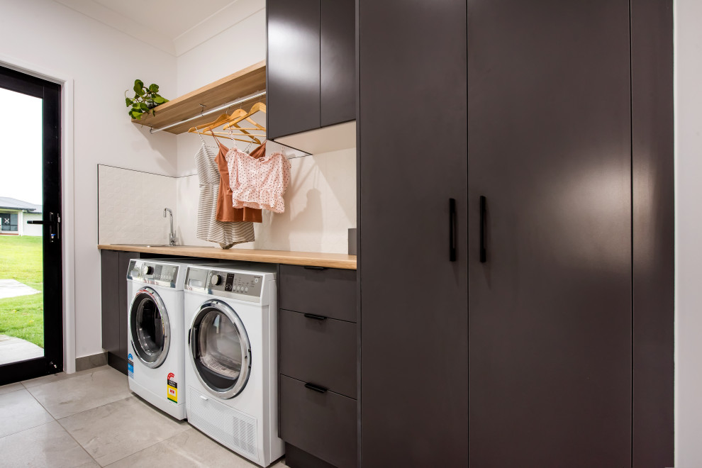 This is an example of a mid-sized contemporary single-wall dedicated laundry room in Other with a drop-in sink, flat-panel cabinets, grey cabinets, wood benchtops, white splashback, ceramic splashback, white walls, ceramic floors, a side-by-side washer and dryer and grey floor.