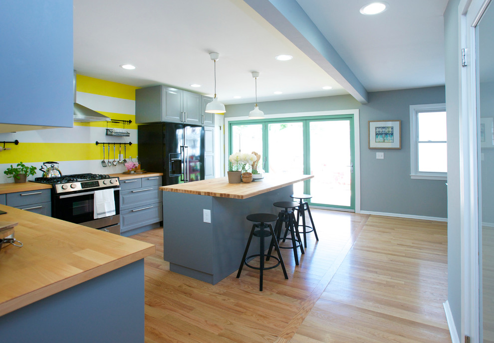 Photo of a contemporary l-shaped eat-in kitchen in New York with a drop-in sink, raised-panel cabinets, grey cabinets, wood benchtops, black appliances, medium hardwood floors and with island.