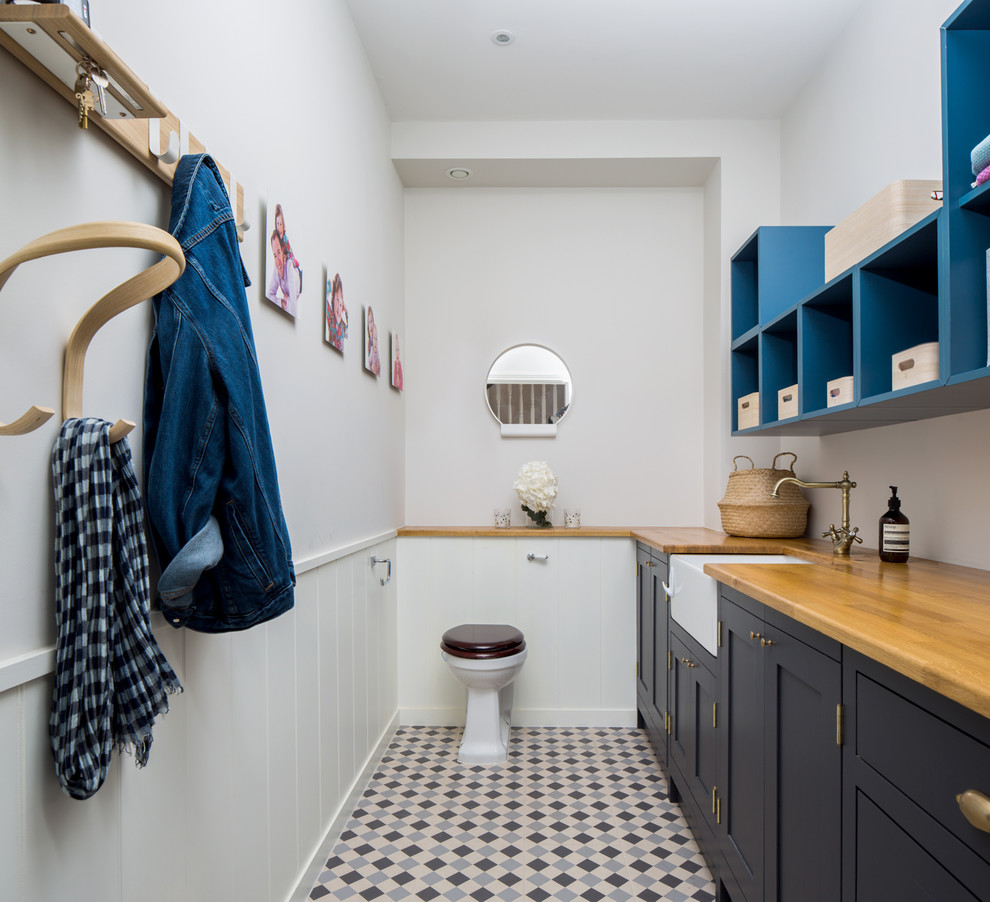 Photo of a large transitional single-wall utility room in London with blue cabinets, grey walls, ceramic floors, a farmhouse sink, wood benchtops, multi-coloured floor, brown benchtop and shaker cabinets.
