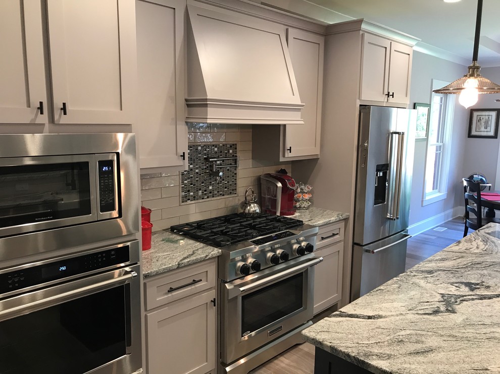 Photo of a mid-sized arts and crafts u-shaped open plan kitchen in Raleigh with a farmhouse sink, shaker cabinets, grey cabinets, granite benchtops, white splashback, porcelain splashback, stainless steel appliances, light hardwood floors, with island and brown floor.