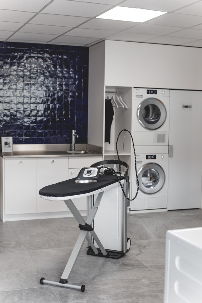 This is an example of a large modern single-wall dedicated laundry room in Other with an integrated sink, white cabinets, stainless steel benchtops, blue splashback, ceramic splashback, blue walls, ceramic floors, a stacked washer and dryer, grey floor and grey benchtop.