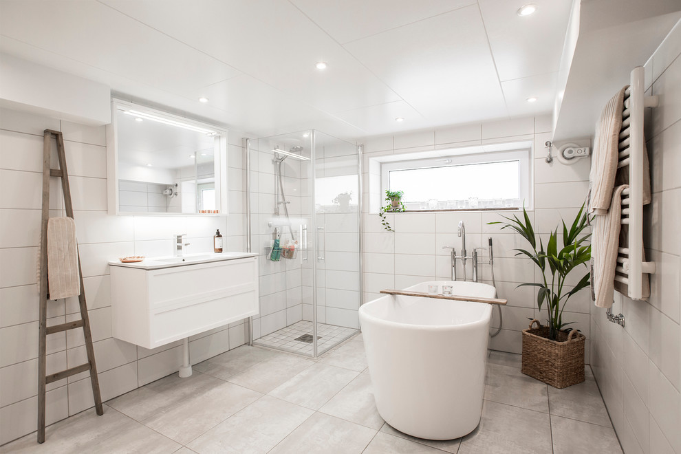 Photo of a mid-sized contemporary master bathroom in Malmo with white cabinets, a freestanding tub, a corner shower, gray tile, white tile, white walls, a console sink, limestone floors and a hinged shower door.