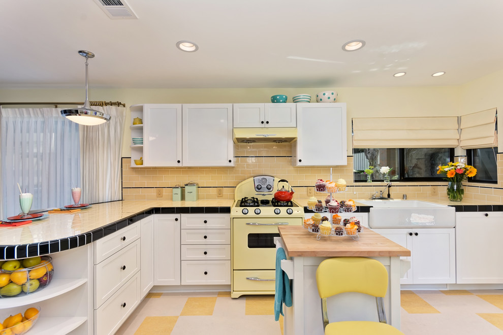 Photo of a mid-sized traditional l-shaped eat-in kitchen in San Diego with a farmhouse sink, shaker cabinets, white cabinets, tile benchtops, yellow splashback, subway tile splashback, coloured appliances, linoleum floors and a peninsula.
