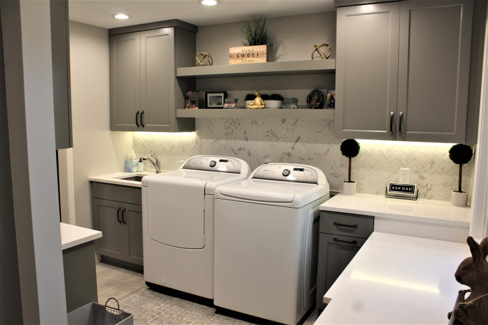 Inspiration for a large transitional u-shaped utility room in Detroit with an undermount sink, flat-panel cabinets, grey cabinets, quartz benchtops, grey splashback, stone tile splashback, grey walls, porcelain floors, a side-by-side washer and dryer, grey floor and white benchtop.