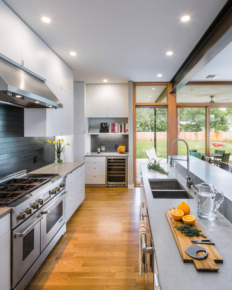 Contemporary open plan kitchen in Austin with a double-bowl sink, flat-panel cabinets, white cabinets, black splashback, stainless steel appliances, medium hardwood floors, with island and metal splashback.