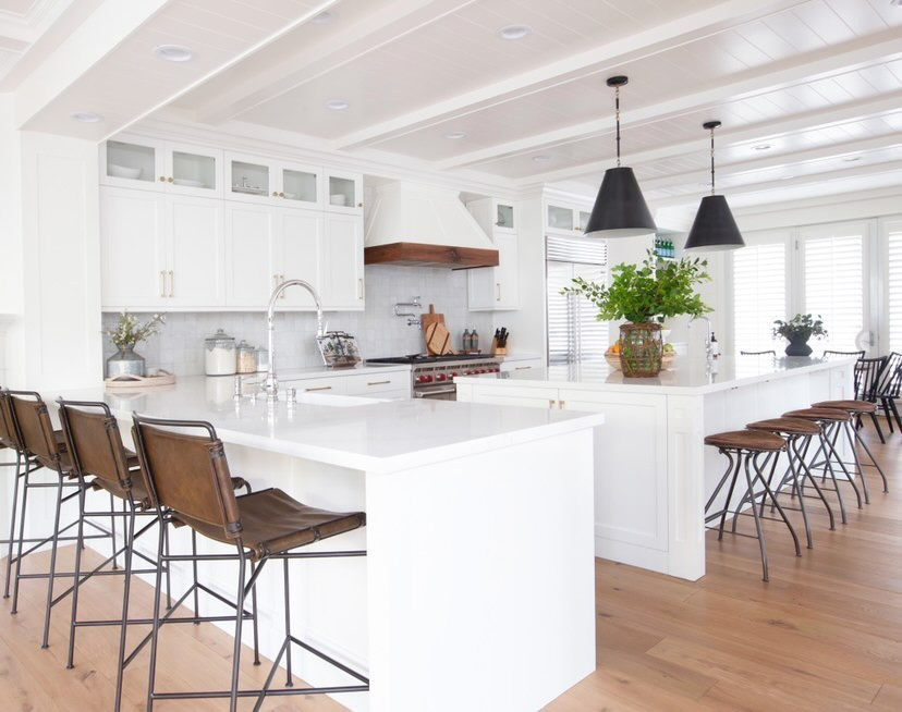 Photo of a l-shaped kitchen in Los Angeles with glass-front cabinets, white cabinets, marble benchtops, white splashback, stainless steel appliances, light hardwood floors, with island, white floor, white benchtop and exposed beam.