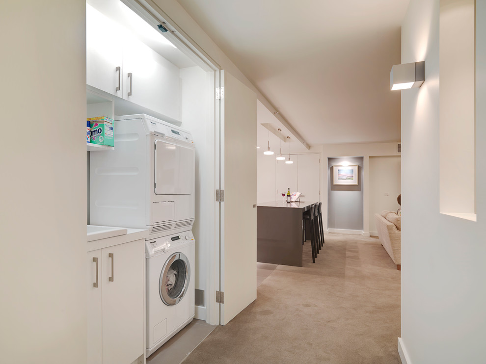 Photo of a small modern single-wall laundry cupboard in Melbourne with a drop-in sink, flat-panel cabinets, white cabinets, quartz benchtops, porcelain floors, a concealed washer and dryer and white walls.