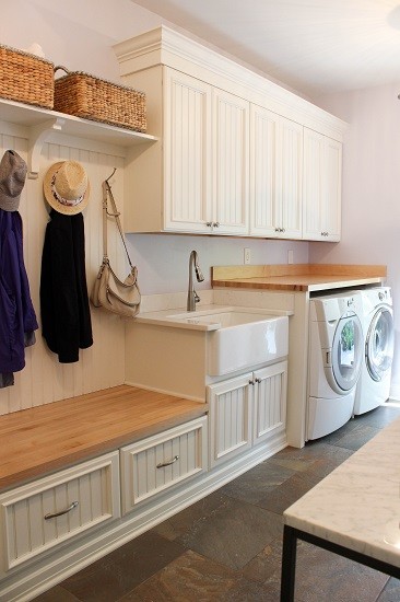 Photo of a mid-sized traditional single-wall utility room in Other with a farmhouse sink, recessed-panel cabinets, white cabinets, wood benchtops, purple walls, porcelain floors, a side-by-side washer and dryer and grey floor.