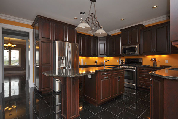 Porcelain tile kitchen photo with a farmhouse sink, raised-panel cabinets, dark wood cabinets, granite countertops, multicolored backsplash, ceramic backsplash, stainless steel appliances and an island