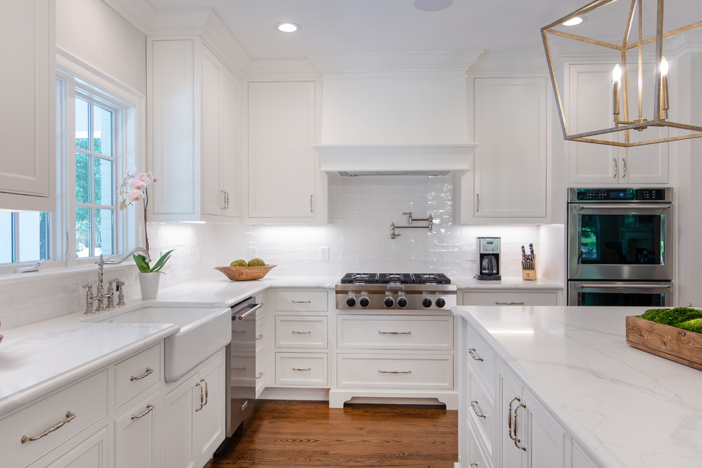 Mid-sized country l-shaped kitchen in Charlotte with a farmhouse sink, beaded inset cabinets, white cabinets, quartzite benchtops, white splashback, subway tile splashback, stainless steel appliances, medium hardwood floors, with island and white benchtop.