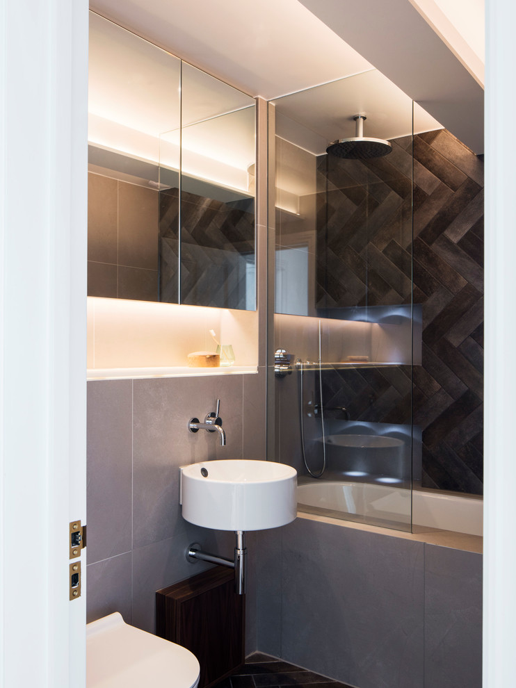 This is an example of a mid-sized contemporary kids bathroom in London with a drop-in tub, a shower/bathtub combo, a wall-mount toilet, gray tile, cement tile, grey walls, porcelain floors, a wall-mount sink, black floor and white benchtops.