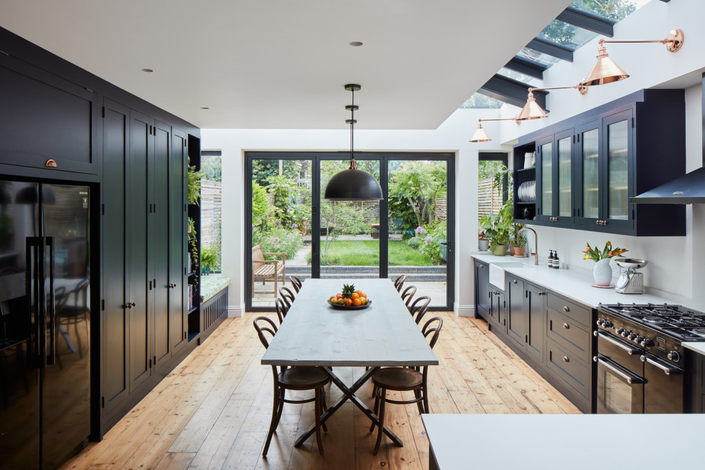 Design ideas for a large eclectic l-shaped eat-in kitchen in London with a farmhouse sink, shaker cabinets, blue cabinets, quartz benchtops, white splashback, engineered quartz splashback, black appliances, light hardwood floors, no island, grey benchtop and exposed beam.