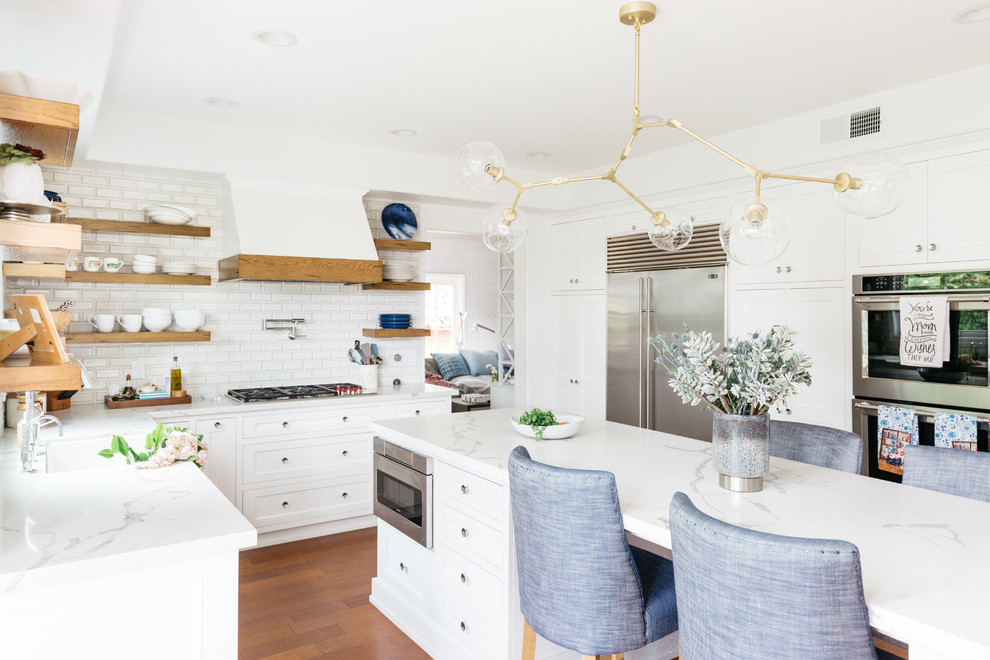 Eat-in kitchen - mid-sized transitional u-shaped medium tone wood floor and brown floor eat-in kitchen idea in Los Angeles with a farmhouse sink, recessed-panel cabinets, white cabinets, quartzite countertops, white backsplash, subway tile backsplash, stainless steel appliances, an island and white countertops