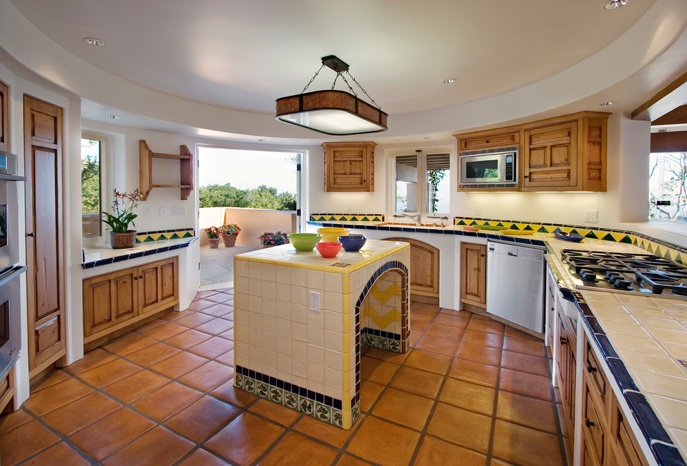 Photo of a mediterranean separate kitchen in Santa Barbara with an undermount sink, recessed-panel cabinets, medium wood cabinets, tile benchtops, multi-coloured splashback, ceramic splashback, stainless steel appliances and terra-cotta floors.