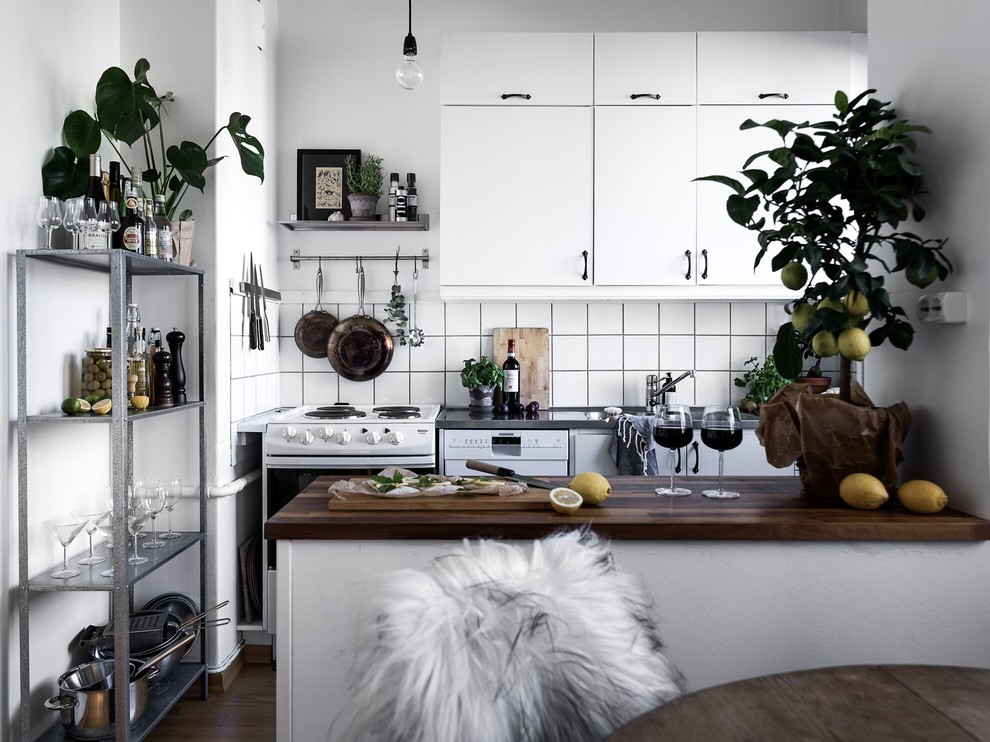 This is an example of a mid-sized scandinavian single-wall kitchen in Stockholm with flat-panel cabinets, white cabinets, white splashback, white appliances, dark hardwood floors, a peninsula, wood benchtops and ceramic splashback.