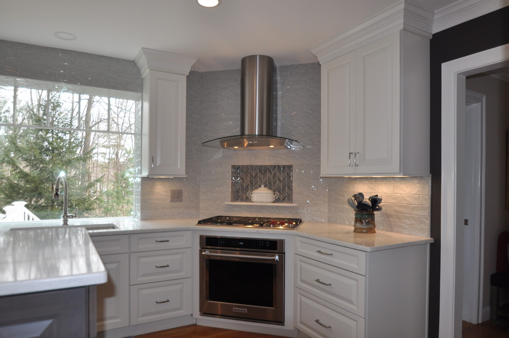 This is an example of a mid-sized transitional l-shaped open plan kitchen in Boston with an undermount sink, raised-panel cabinets, white cabinets, quartz benchtops, grey splashback, porcelain splashback, stainless steel appliances, medium hardwood floors, with island, brown floor and white benchtop.