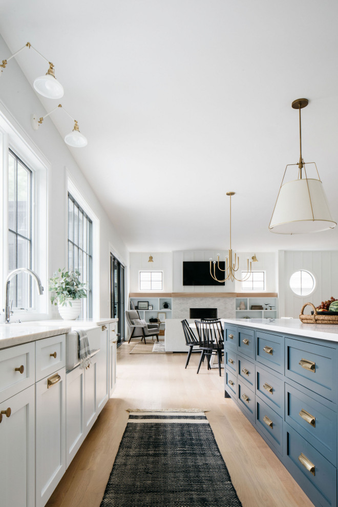 This is an example of a coastal open plan kitchen in Chicago with a belfast sink, shaker cabinets, blue cabinets, white splashback, light hardwood flooring, an island and white worktops.