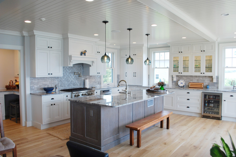 Photo of a large beach style u-shaped open plan kitchen in Providence with a farmhouse sink, shaker cabinets, white cabinets, quartz benchtops, grey splashback, subway tile splashback, stainless steel appliances, light hardwood floors, with island, grey benchtop and timber.
