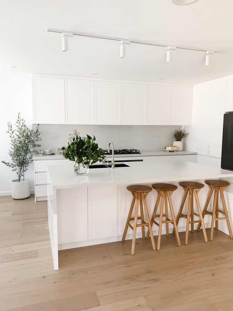 Inspiration for a mid-sized eat-in kitchen in Melbourne with an undermount sink, shaker cabinets, white cabinets, quartz benchtops, engineered quartz splashback, black appliances, light hardwood floors and with island.