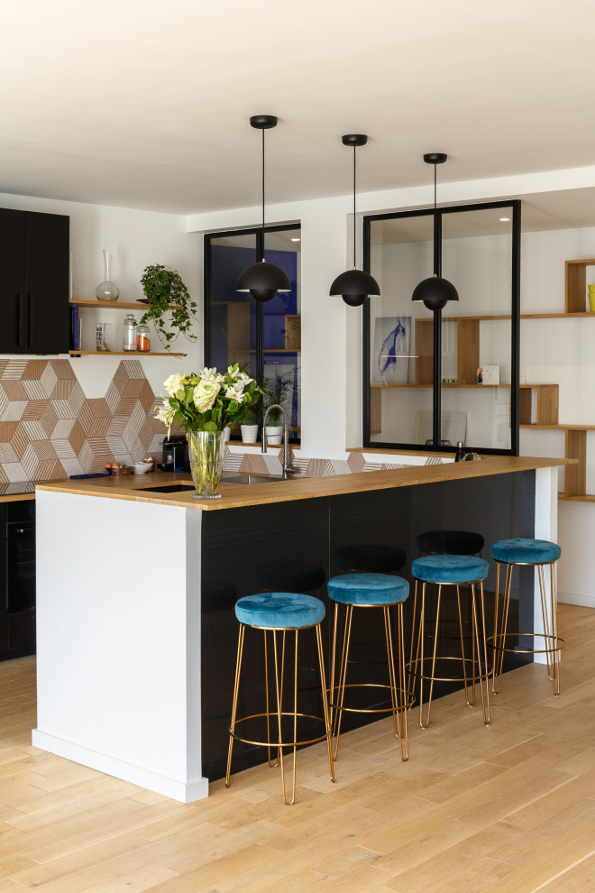 This is an example of a mid-sized modern galley open plan kitchen in Paris with a single-bowl sink, flat-panel cabinets, black cabinets, wood benchtops, orange splashback, ceramic splashback, black appliances, light hardwood floors, with island, beige floor and beige benchtop.