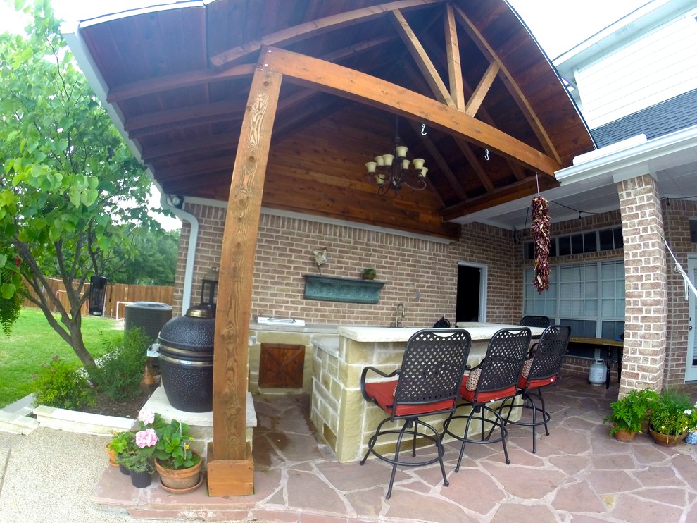 This is an example of a mid-sized country backyard patio in Dallas with an outdoor kitchen, natural stone pavers and a roof extension.