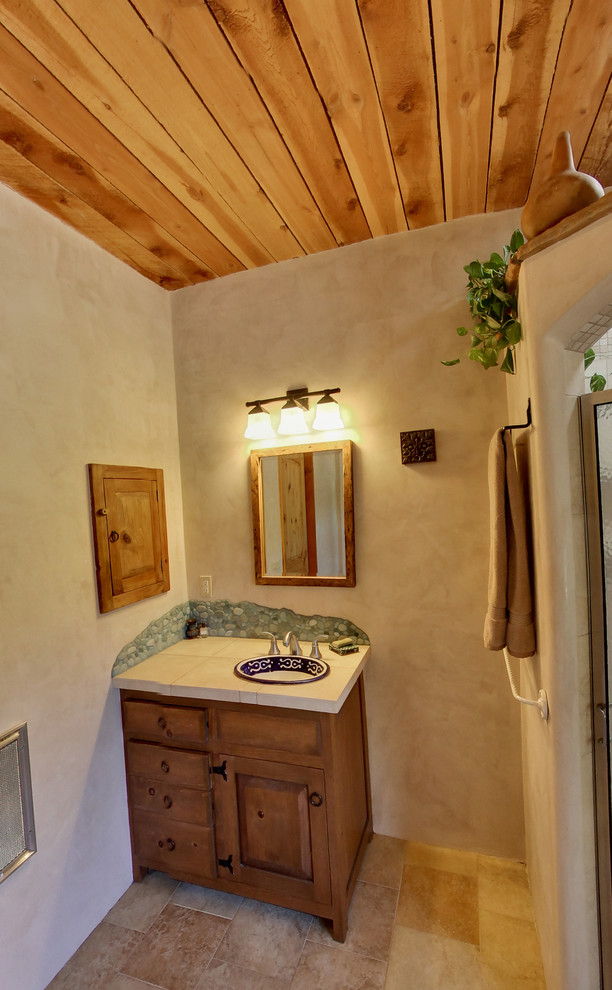 3/4 bathroom with pebble tile, brown walls, travertine floors, a drop-in sink, tile benchtops and beige floor.
