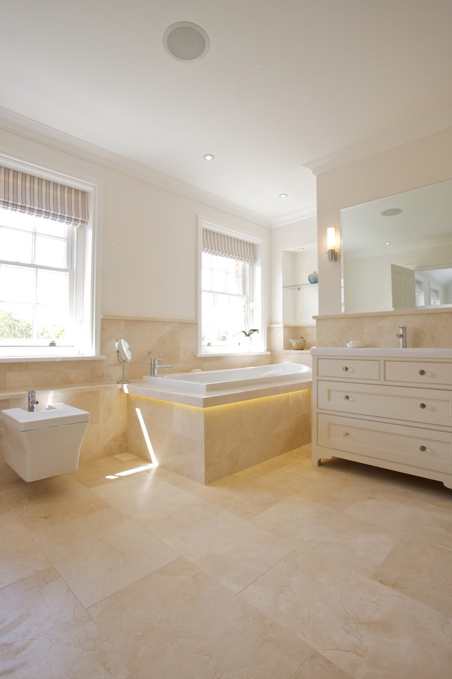 Traditional bathroom in Wiltshire with beige tile, stone tile, beige walls, marble floors, a console sink, beige cabinets, a drop-in tub, a bidet and beaded inset cabinets.