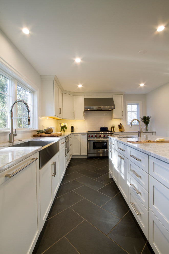 Photo of a large transitional l-shaped kitchen in Charleston with a farmhouse sink, shaker cabinets, white cabinets, quartz benchtops, white splashback, porcelain splashback, stainless steel appliances, slate floors, with island, black floor and white benchtop.