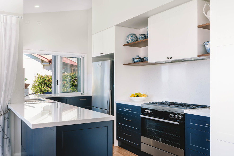 Inspiration for a transitional u-shaped kitchen in Sunshine Coast with a double-bowl sink, shaker cabinets, blue cabinets, white splashback, stainless steel appliances, light hardwood floors, a peninsula and white benchtop.