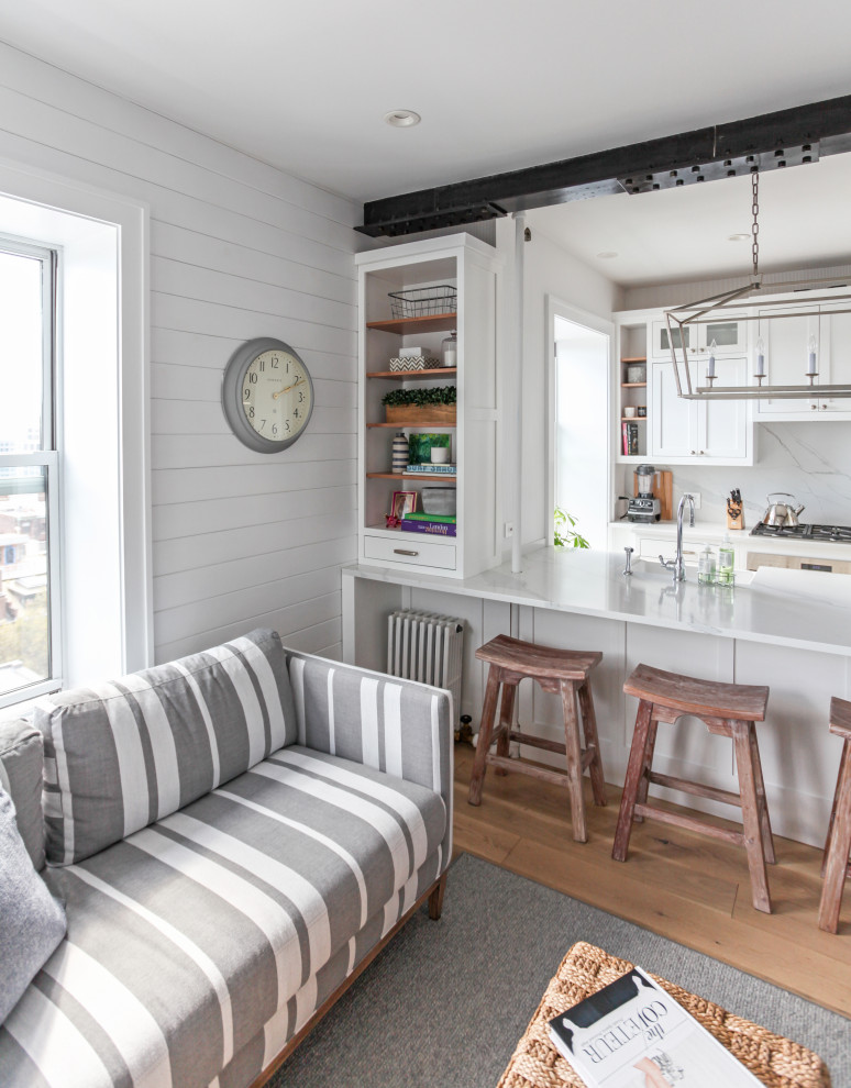 Small beach style single-wall eat-in kitchen in New York with a farmhouse sink, recessed-panel cabinets, white cabinets, quartz benchtops, white splashback, engineered quartz splashback, stainless steel appliances, light hardwood floors, a peninsula and white benchtop.