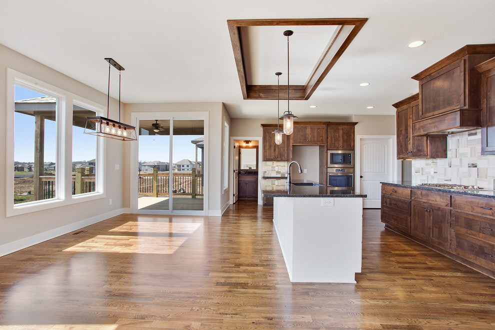 This is an example of a country eat-in kitchen in Kansas City with an undermount sink, shaker cabinets, brown cabinets, granite benchtops, multi-coloured splashback, ceramic splashback, stainless steel appliances, medium hardwood floors, with island, brown floor and black benchtop.