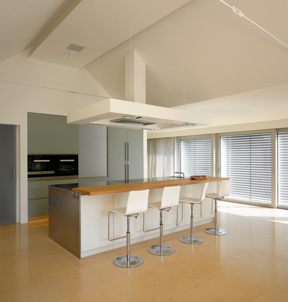 This is an example of a large contemporary open plan kitchen in London with flat-panel cabinets, white cabinets, stainless steel benchtops, with island and beige floor.