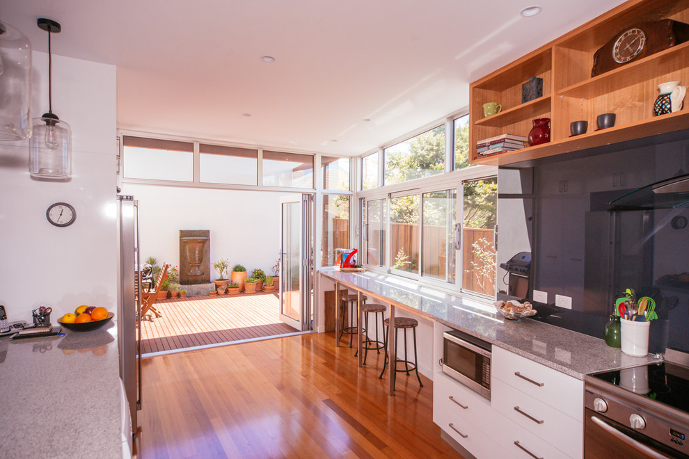 Contemporary galley eat-in kitchen in Hobart with a double-bowl sink, flat-panel cabinets, white cabinets, granite benchtops, black splashback, glass sheet splashback, stainless steel appliances, medium hardwood floors and no island.