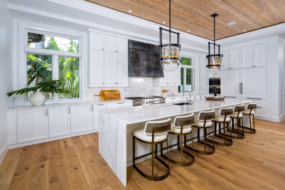 Photo of a beach style l-shaped kitchen in Miami with an undermount sink, recessed-panel cabinets, white cabinets, white splashback, stainless steel appliances, medium hardwood floors, with island, brown floor, white benchtop and wood.