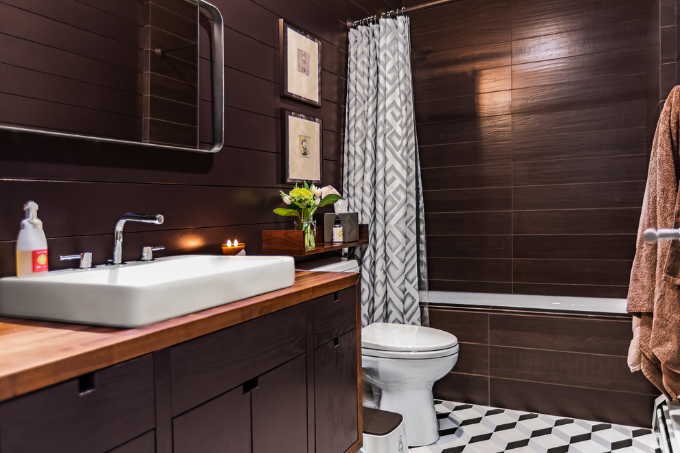 Photo of a mid-sized modern 3/4 bathroom in New York with flat-panel cabinets, dark wood cabinets, a drop-in tub, a shower/bathtub combo, brown tile, brown walls, a vessel sink, wood benchtops, multi-coloured floor, a shower curtain, brown benchtops, a two-piece toilet and ceramic floors.